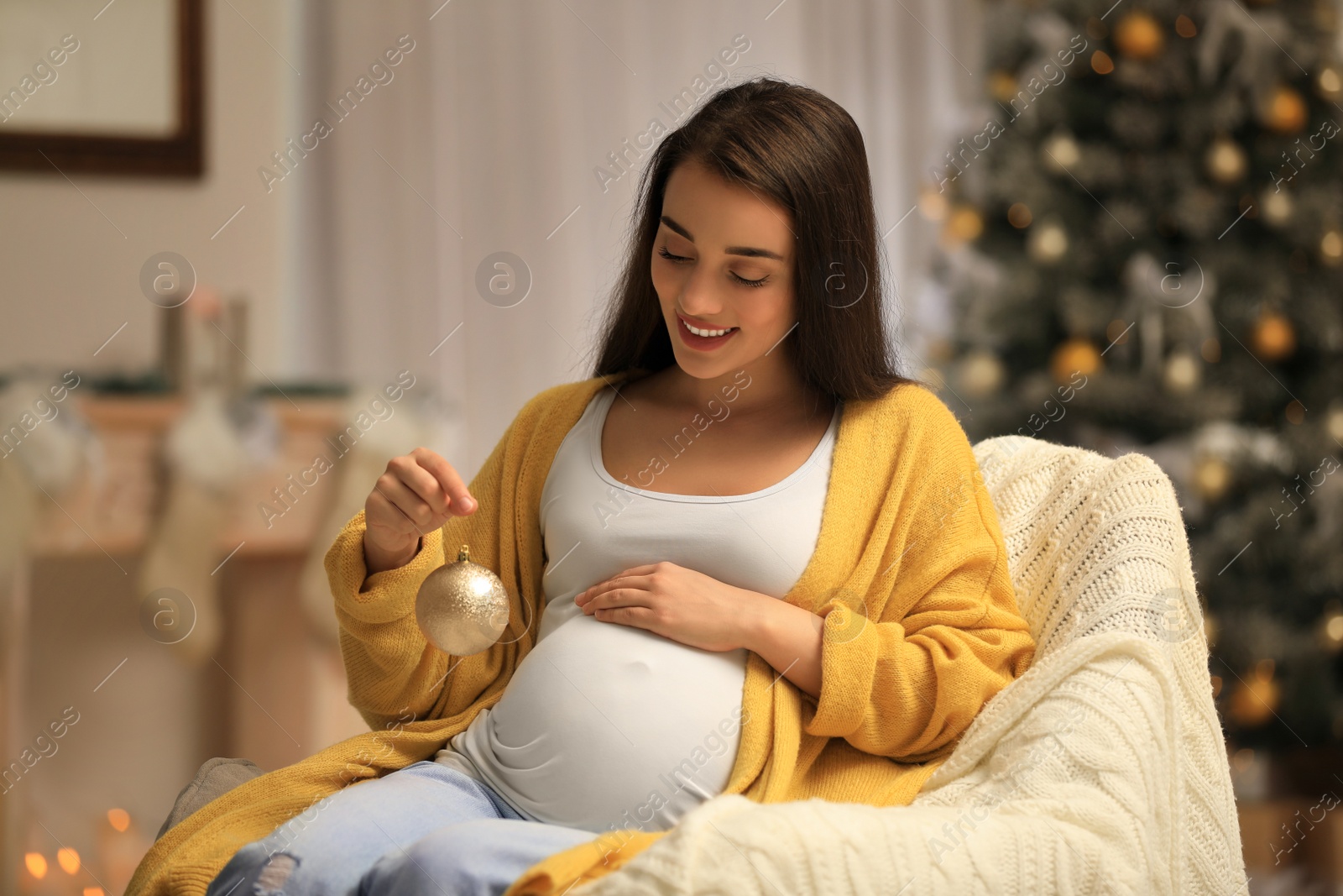 Photo of Happy pregnant woman with Christmas ball in armchair at home. Expecting baby