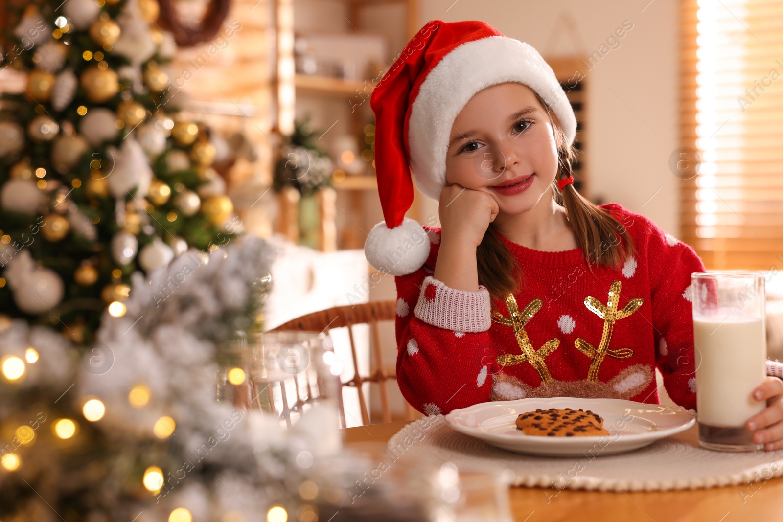 Photo of Cute little girl in Santa hat with delicious Christmas cookie and milk at home, space for text