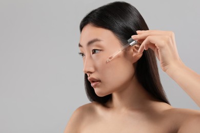 Photo of Beautiful young woman applying cosmetic serum onto her face on grey background, space for text