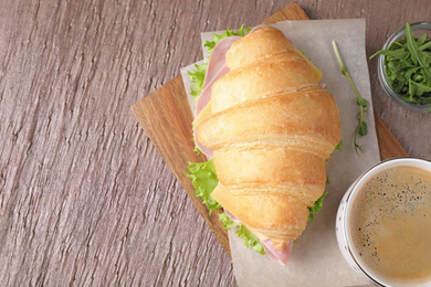 Photo of Tasty croissant sandwich with ham and cup of coffee on brown table, flat lay