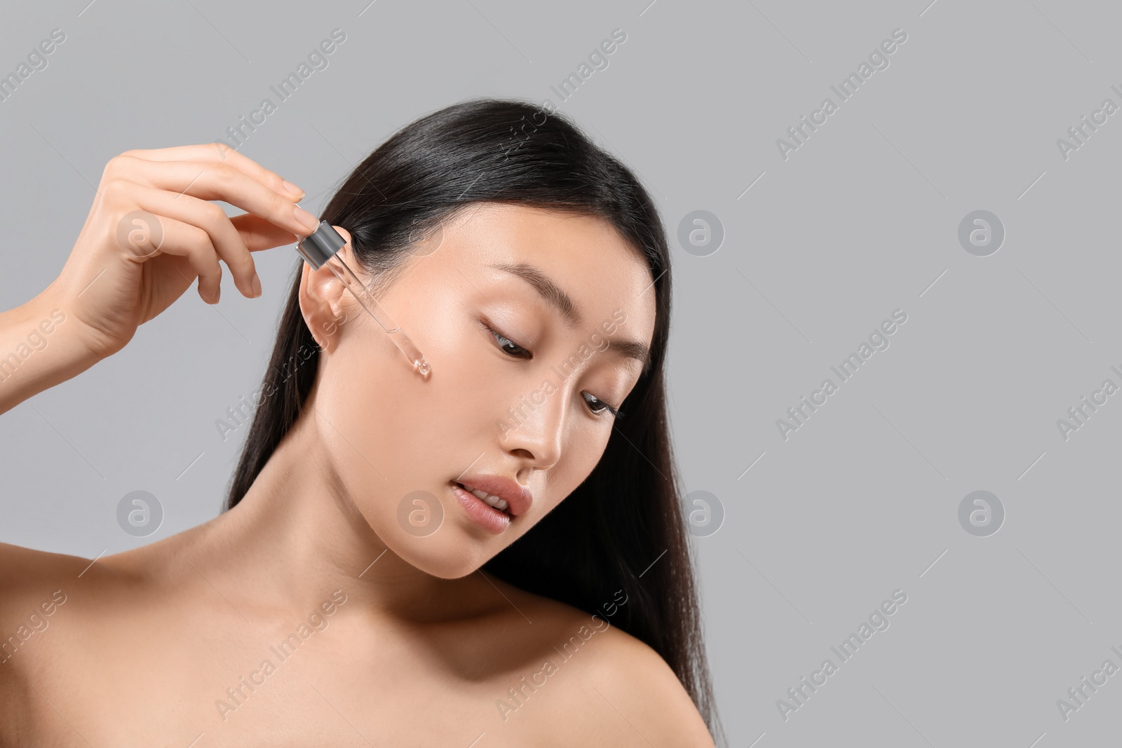 Photo of Beautiful young woman applying cosmetic serum onto her face on grey background, space for text