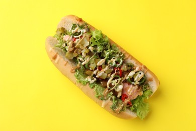 Photo of Delicious hot dog with chili peppers, lettuce, pickles and sauces on yellow background, top view
