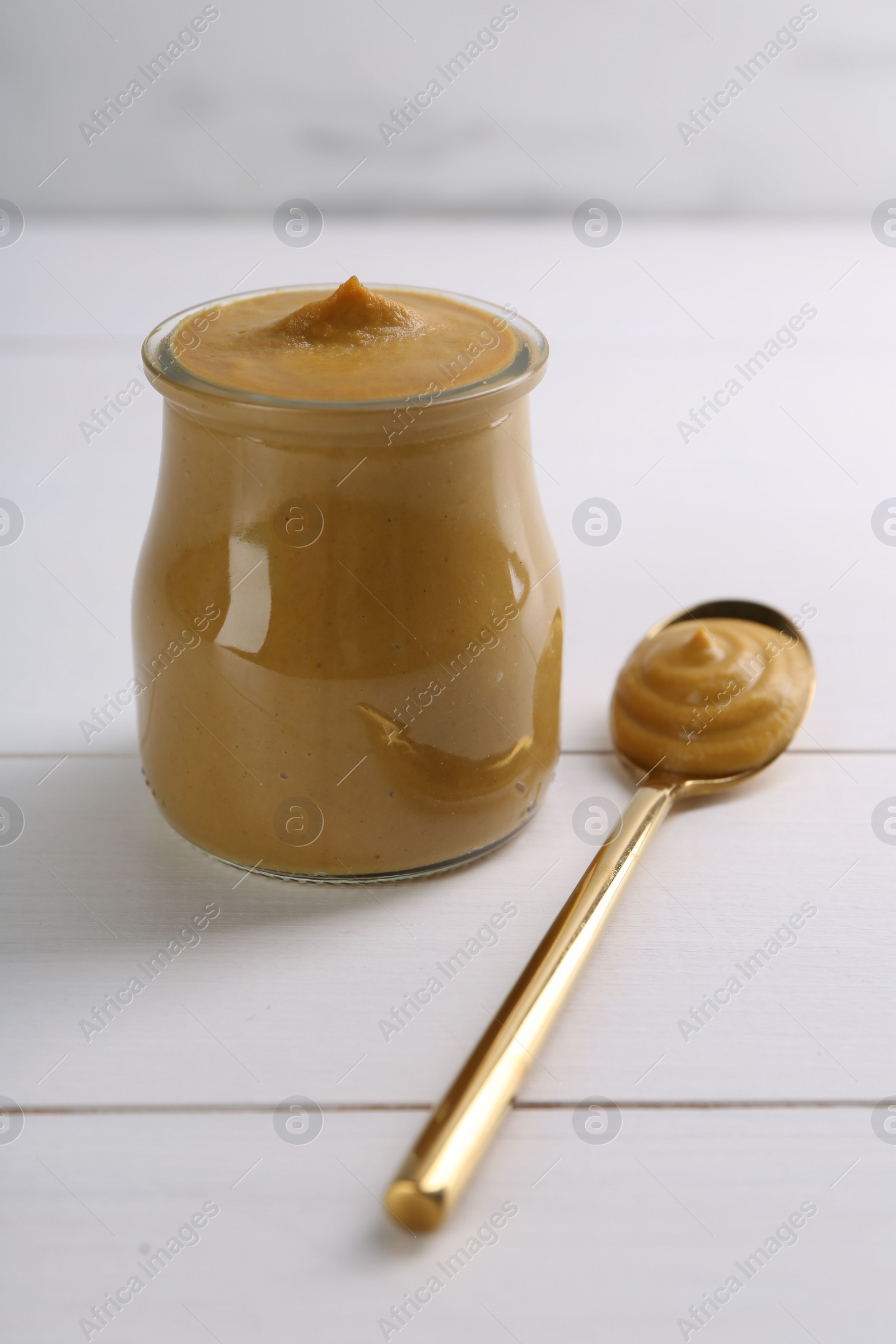 Photo of Spoon and glass jar of tasty mustard sauce on white wooden table, closeup