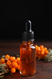 Photo of Ripe sea buckthorn and bottle of essential oil on wooden table against black background
