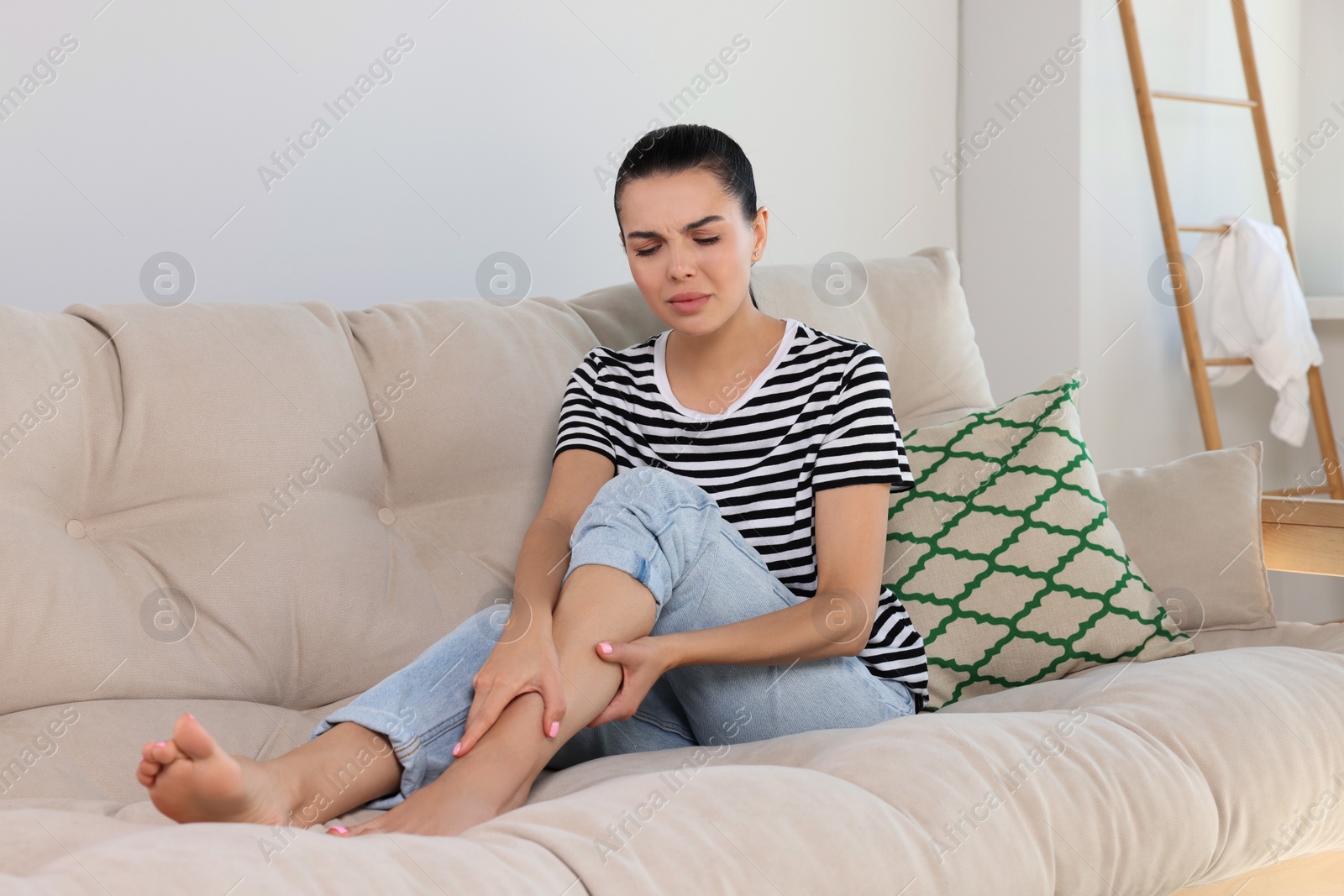 Photo of Young woman rubbing sore leg on sofa at home