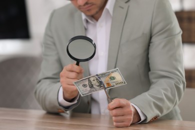 Photo of Expert authenticating 100 dollar banknote with magnifying glass at table in office, closeup. Fake money concept