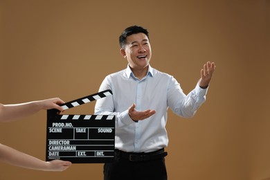 Photo of Happy asian actor performing while second assistant camera holding clapperboard on brown background. Film industry