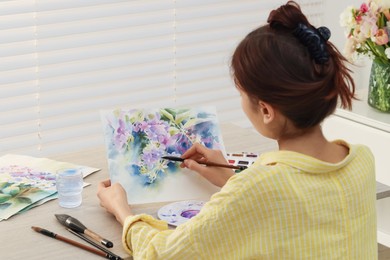 Photo of Woman painting flowers with watercolor at white wooden table indoors, back view. Creative artwork
