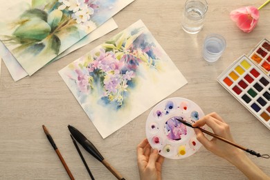 Photo of Woman painting flowers with watercolor at white wooden table, top view. Creative artwork
