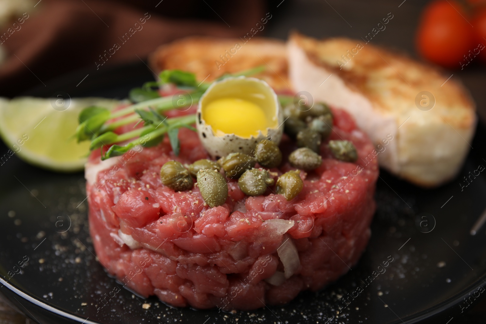 Photo of Tasty beef steak tartare served with quail egg and other accompaniments on black plate, closeup