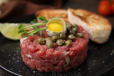 Photo of Tasty beef steak tartare served with quail egg and other accompaniments on black plate, closeup