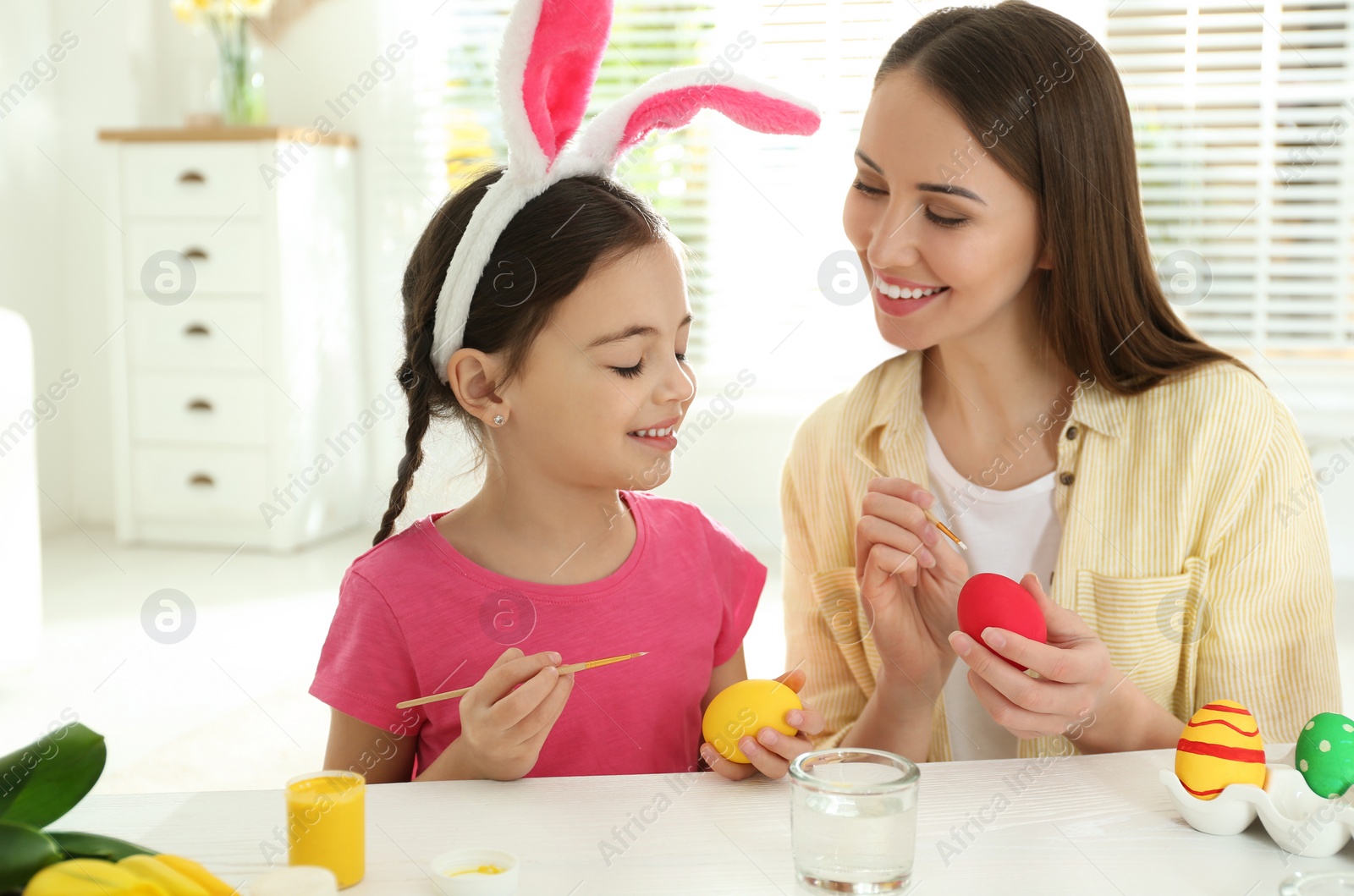 Photo of Happy daughter with bunny ears headband and her mother painting Easter eggs at home