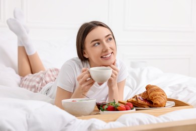 Smiling woman drinking coffee near tray with breakfast on bed