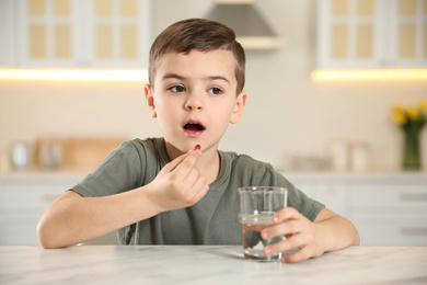 Photo of Little boy with glass of water taking vitamin capsule in kitchen