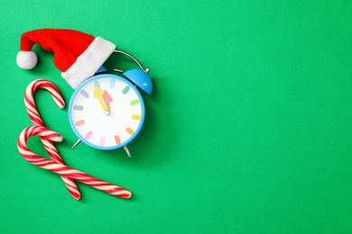 Photo of Alarm clock in Santa hat and candy canes on green background, flat lay with space for text. New Year countdown