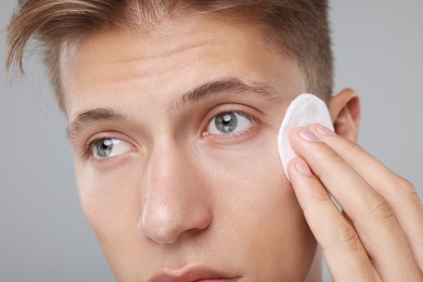 Man cleaning face with cotton pad on grey background, closeup