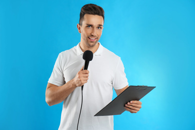 Young male journalist with microphone and clipboard on blue background