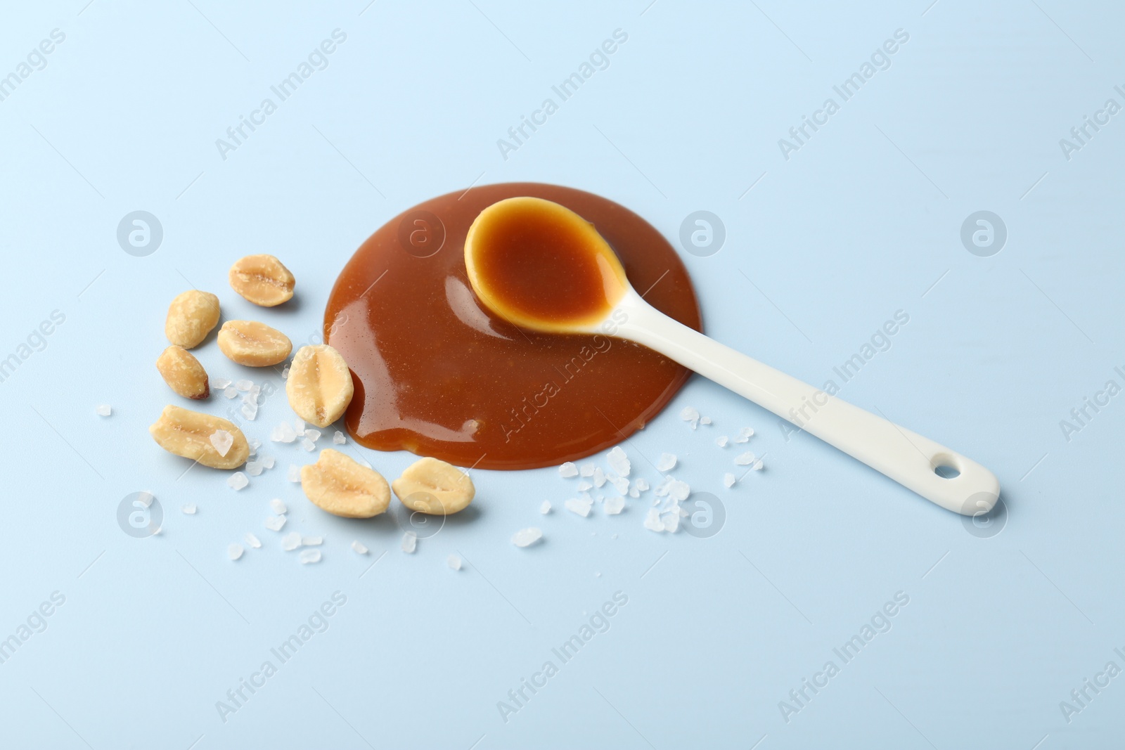 Photo of Spoon with tasty salted caramel, peanuts and salt on light blue background, above view