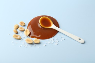 Spoon with tasty salted caramel, peanuts and salt on light blue background, above view