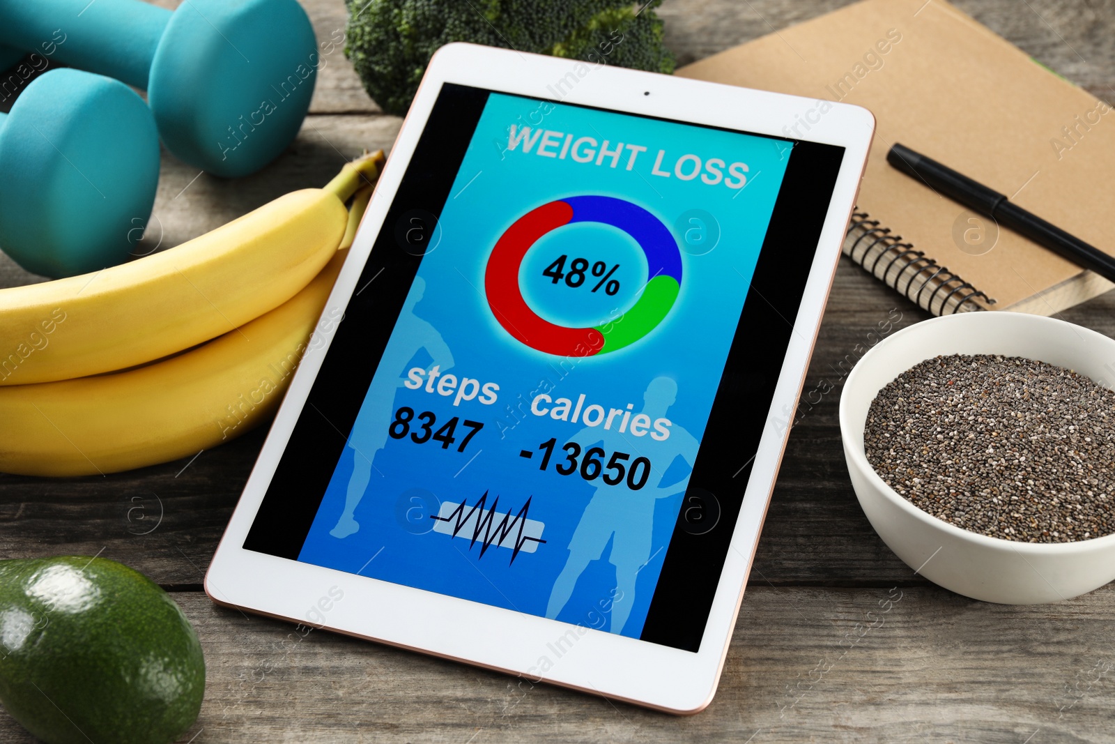 Photo of Tablet with weight loss calculator application, dumbbells and products on wooden table
