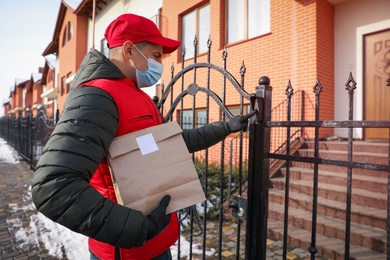 Courier in medical mask holding paper bag with takeaway food and ringing gate bell outdoors. Delivery service during quarantine due to Covid-19 outbreak