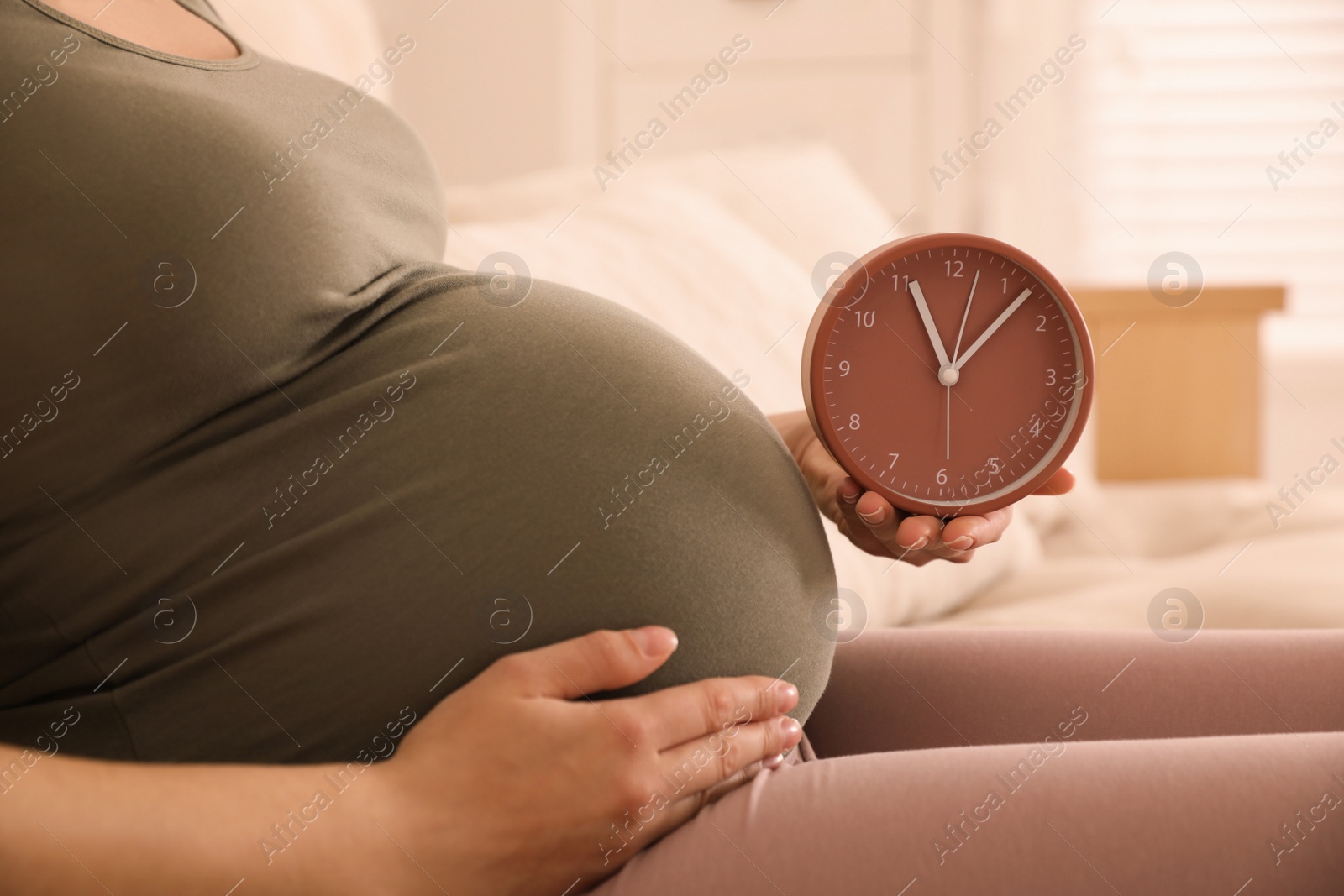 Photo of Young pregnant woman holding clock near her belly at home, closeup. Time to give birth