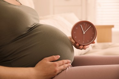 Young pregnant woman holding clock near her belly at home, closeup. Time to give birth