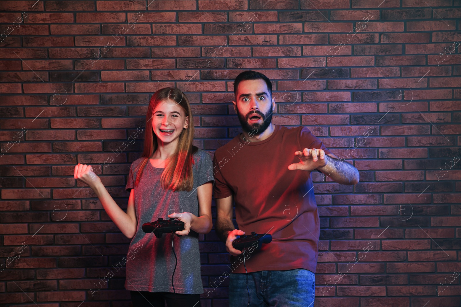 Photo of Young man and teenage girl playing video games with controllers near brick wall