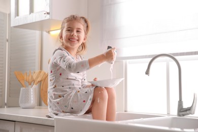 Photo of Naughty girl put her legs into sink while washing dishes in kitchen at home