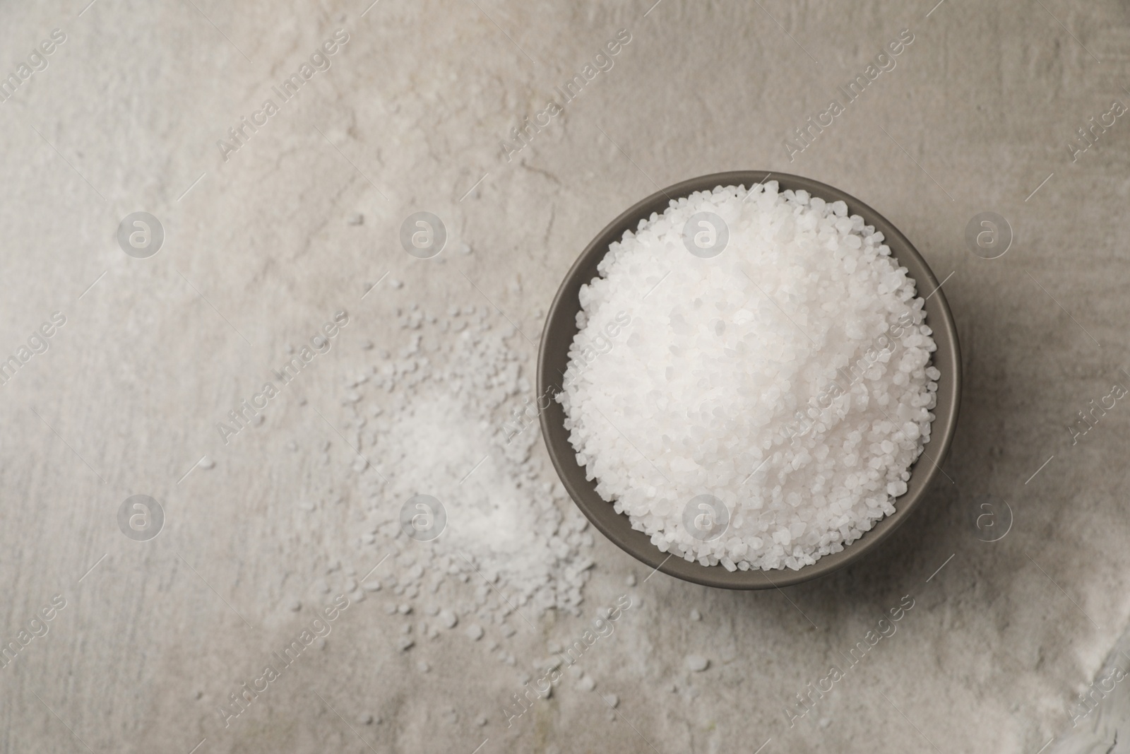 Photo of Natural salt in bowl on grey textured table, top view. Space for text