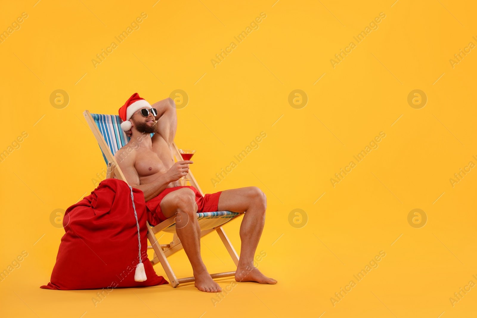 Photo of Muscular young man in Santa hat with deck chair, bag, sunglasses and cocktail on orange background, space for text