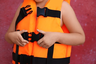 Photo of Little child putting on orange life vest near red wall, closeup