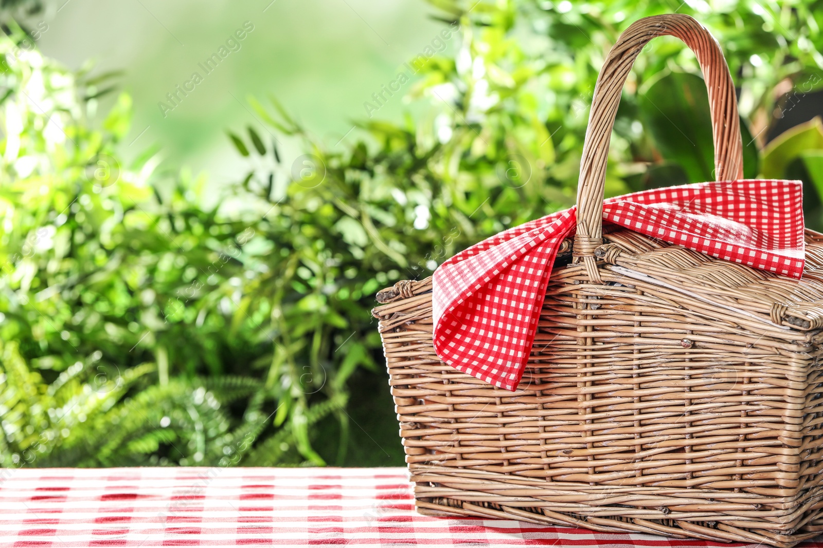 Photo of Closed wicker picnic basket on checkered tablecloth against blurred background, space for text