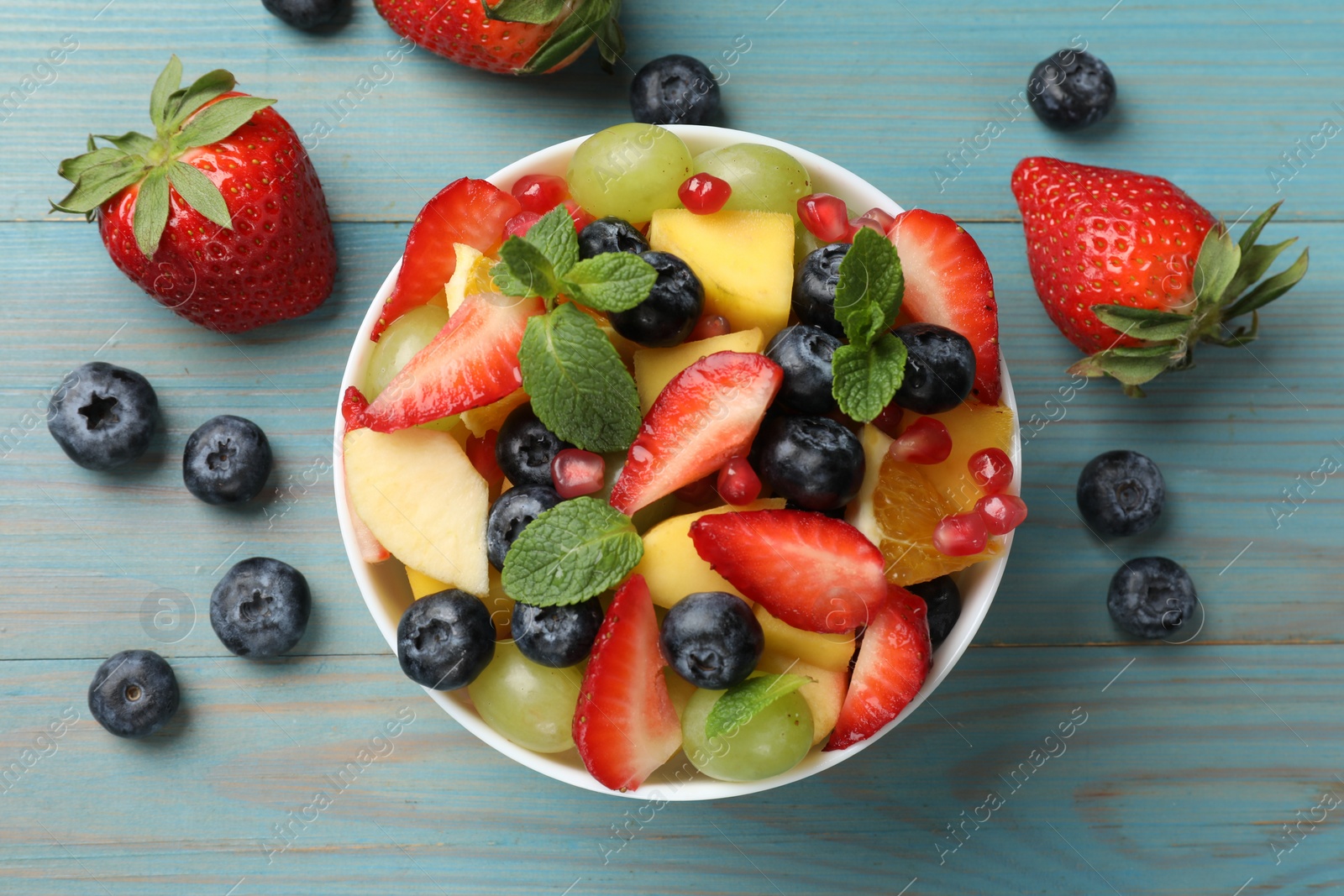Photo of Tasty fruit salad in bowl and ingredients on light blue wooden table, flat lay