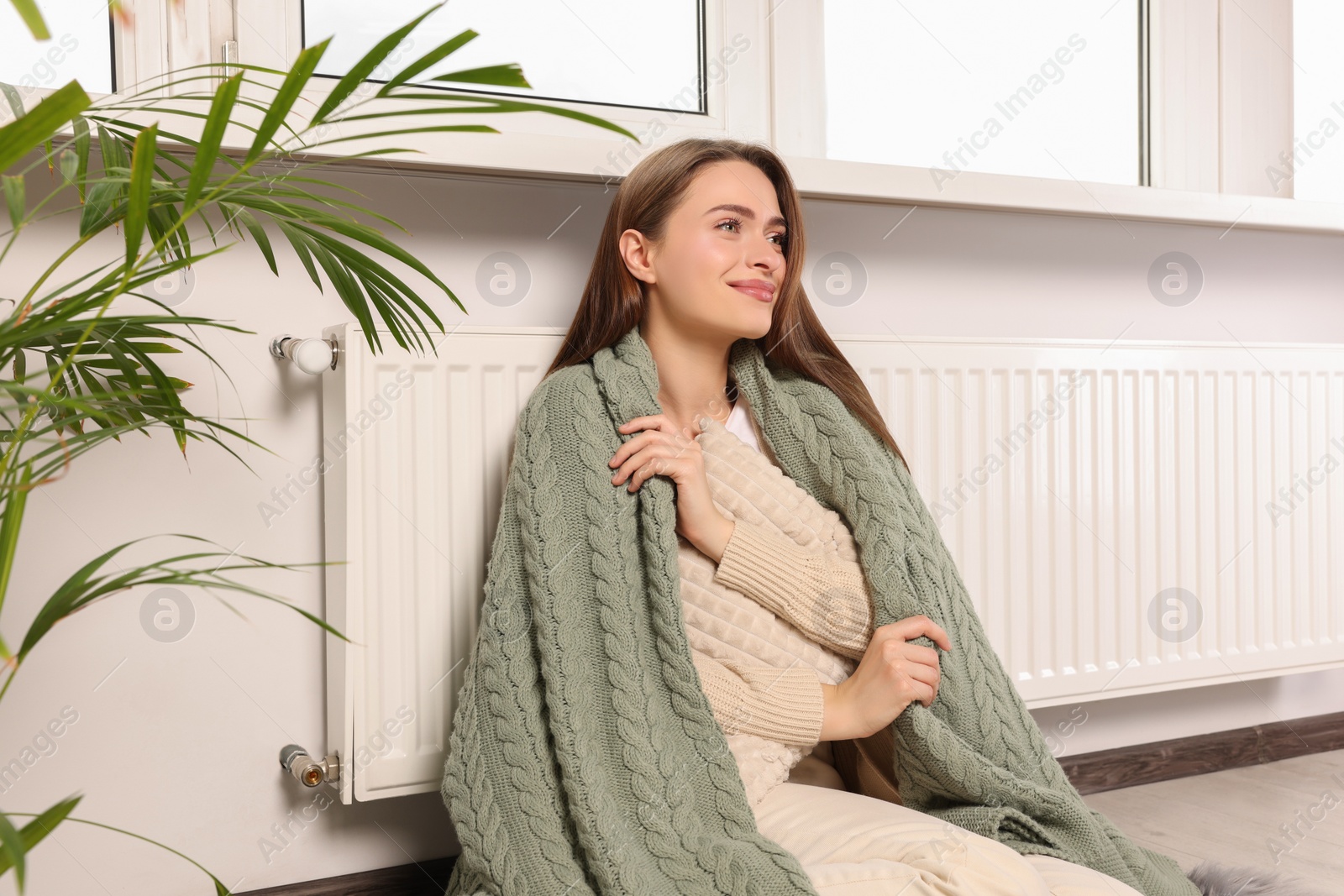 Photo of Woman with blanket hugging pillow near heating radiator indoors