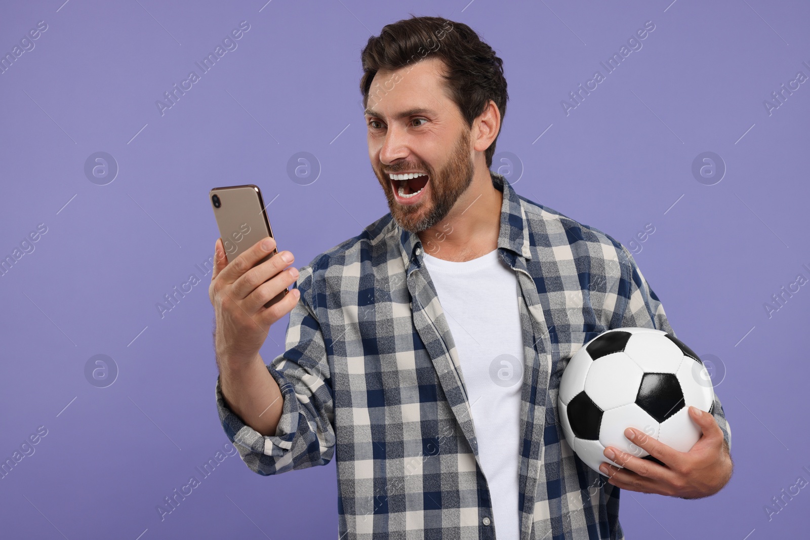 Photo of Emotional sports fan with soccer ball and smartphone on purple background