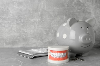 Photo of Educational dental typodont model, piggy bank, coins and calculator on grey table, space for text. Expensive treatment