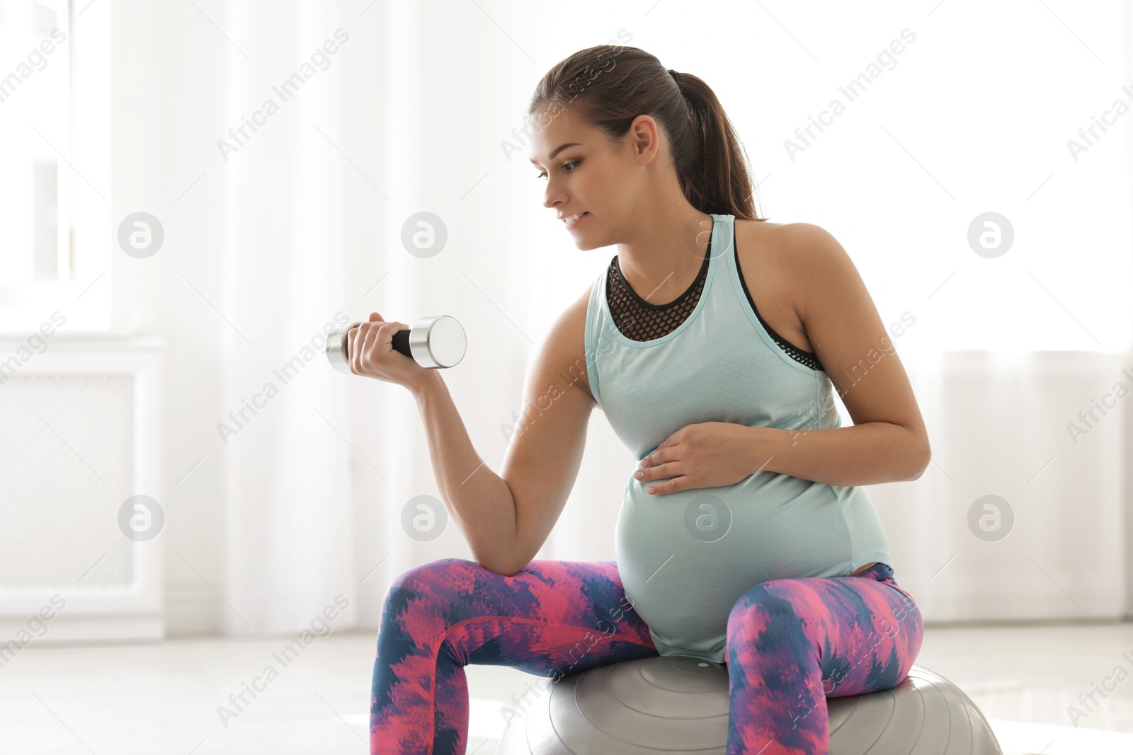 Photo of Young pregnant woman in fitness clothes lifting dumbbell at home