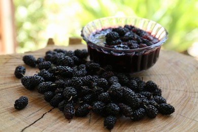 Photo of Delicious ripe black mulberries and bowl of sweet jam on wood stump outdoors