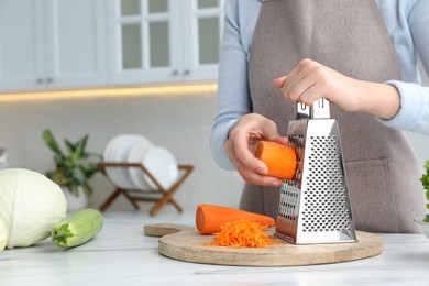 Photo of Woman grating fresh ripe carrot at kitchen table, closeup