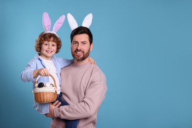 Photo of Happy father and son in cute bunny ears headbands on light blue background. Boy holding Easter basket with painted eggs, space for text