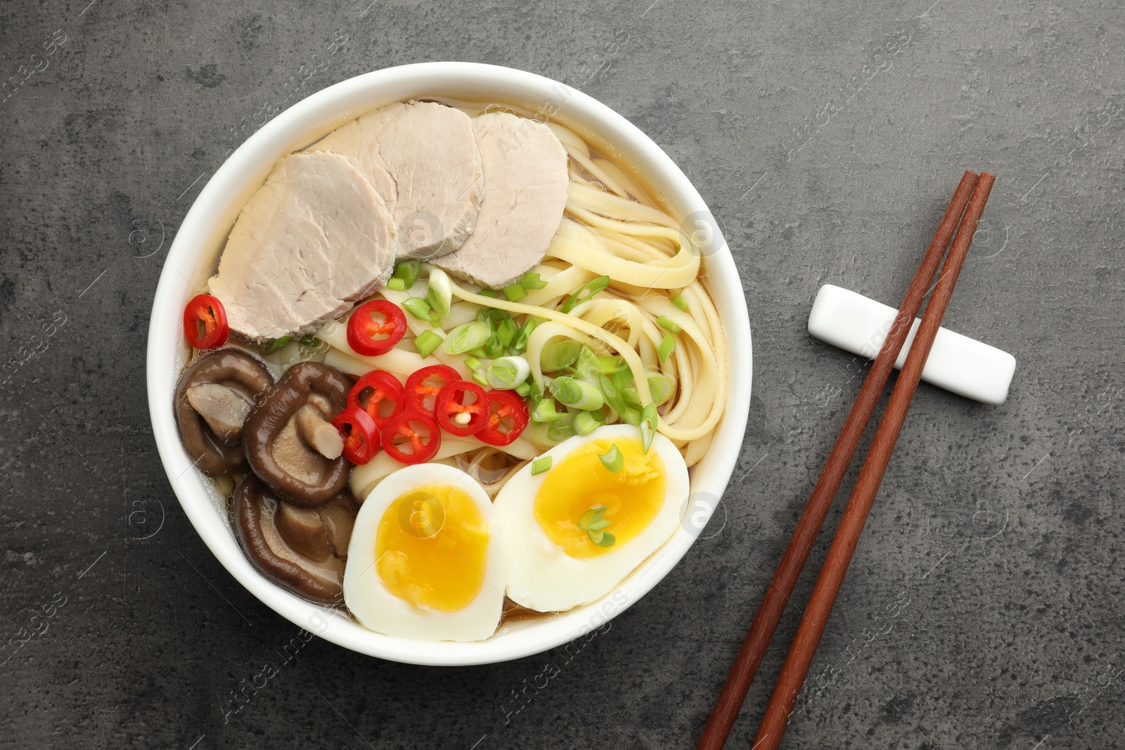 Photo of Delicious ramen with meat in bowl and chopsticks on grey textured table, top view. Noodle soup