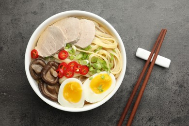 Delicious ramen with meat in bowl and chopsticks on grey textured table, top view. Noodle soup