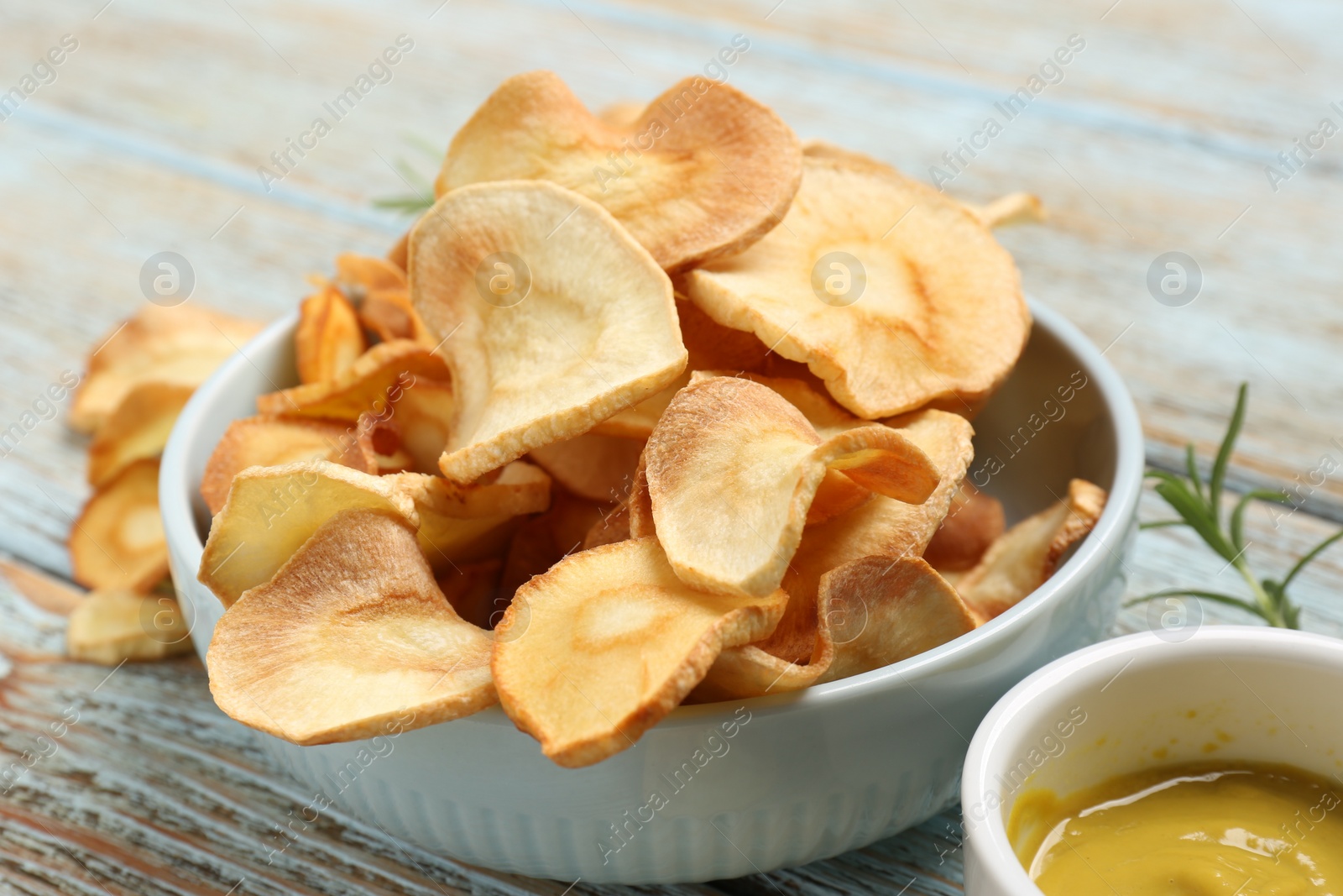 Photo of Tasty homemade parsnip chips with sauce and rosemary on old light blue wooden table, closeup