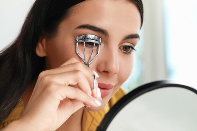 Beautiful young woman using eyelash curler in front of mirror indoors, closeup