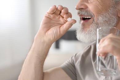 Senior man with glass of water taking pill indoors, closeup