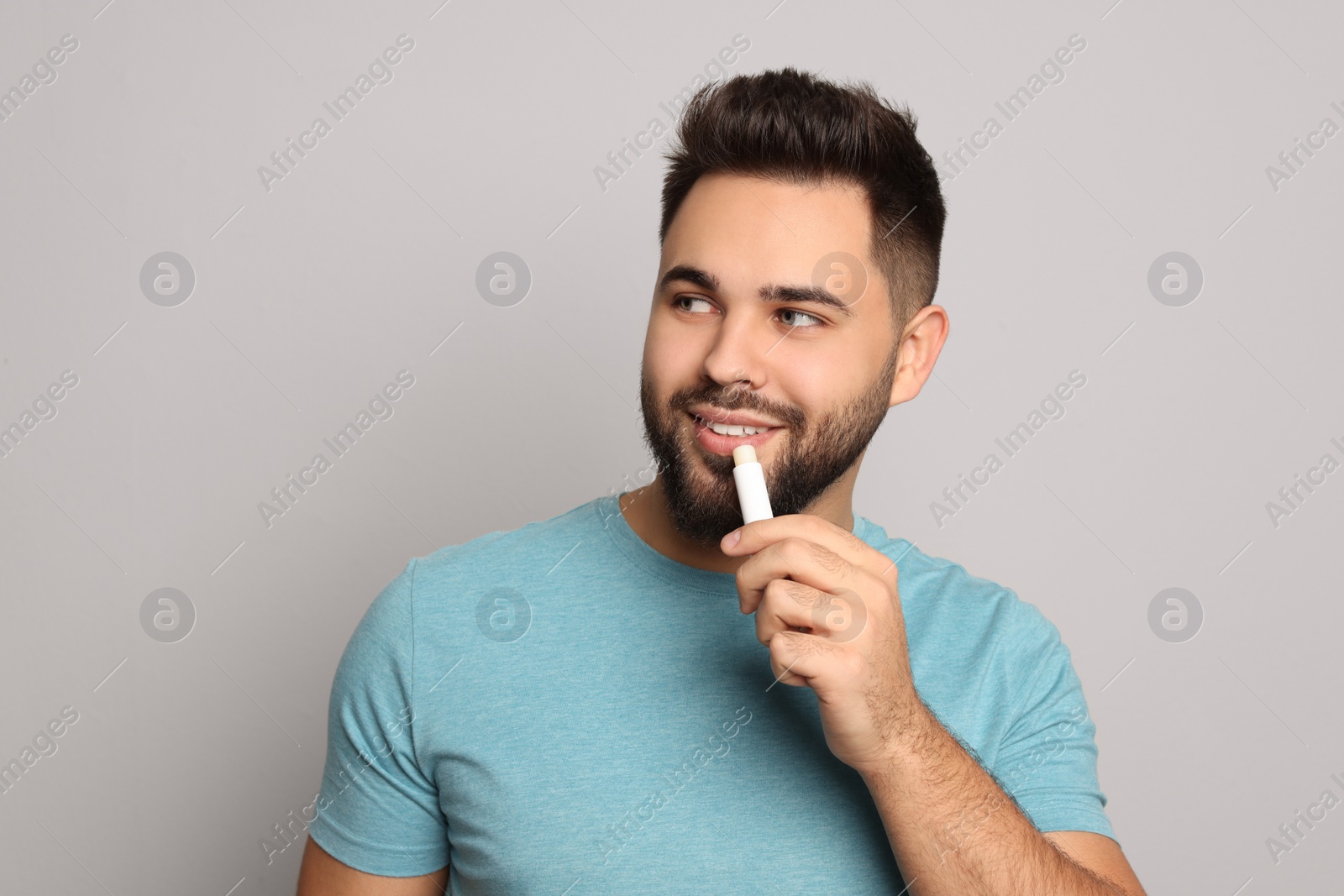 Photo of Young man applying lip balm on grey background, space for text