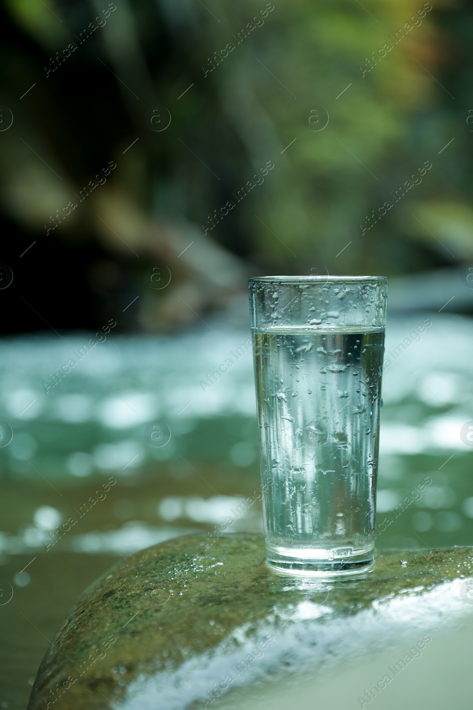 Photo of Glass of fresh water on stone near stream. Space for text