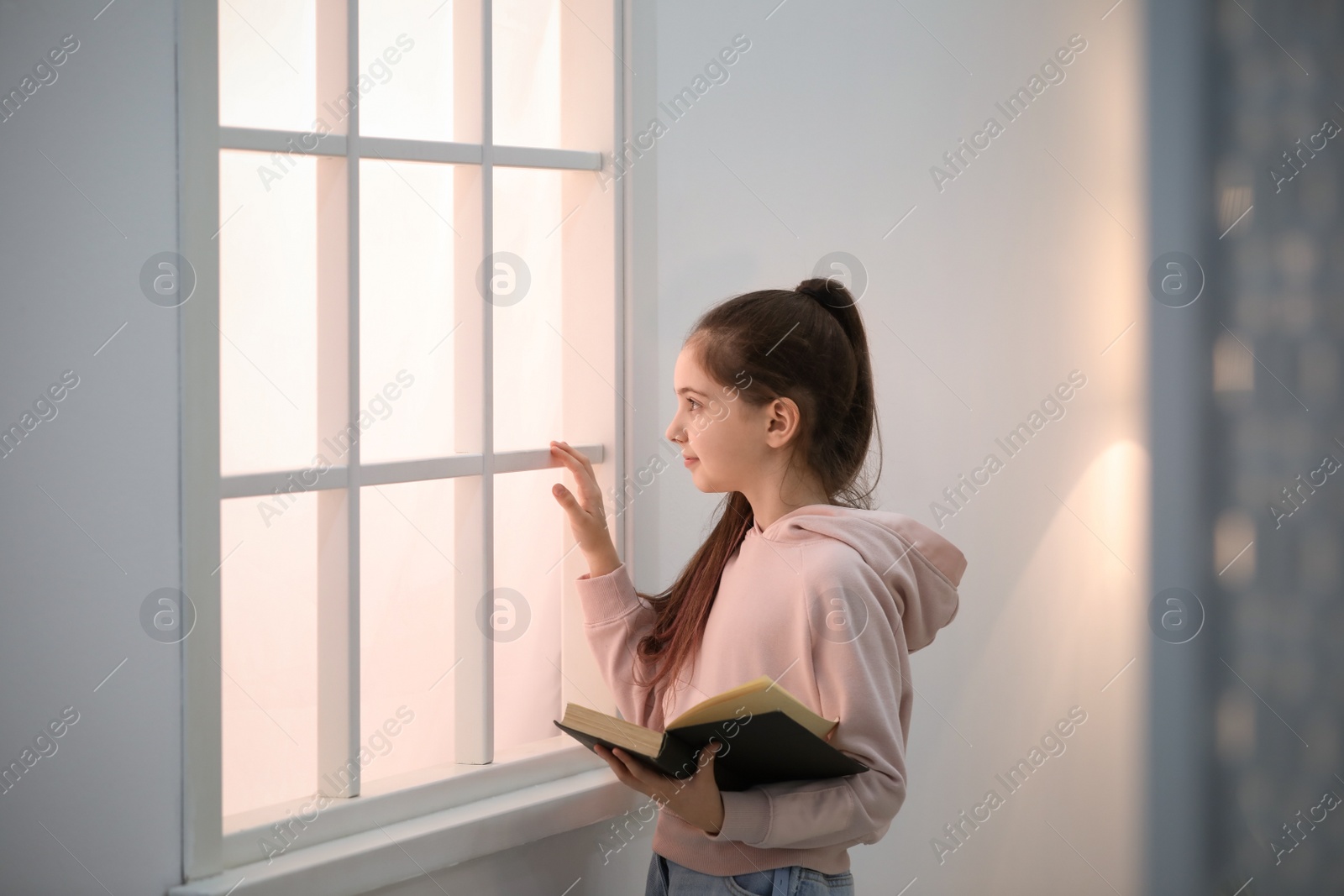 Photo of Cute little girl reading book near window at home