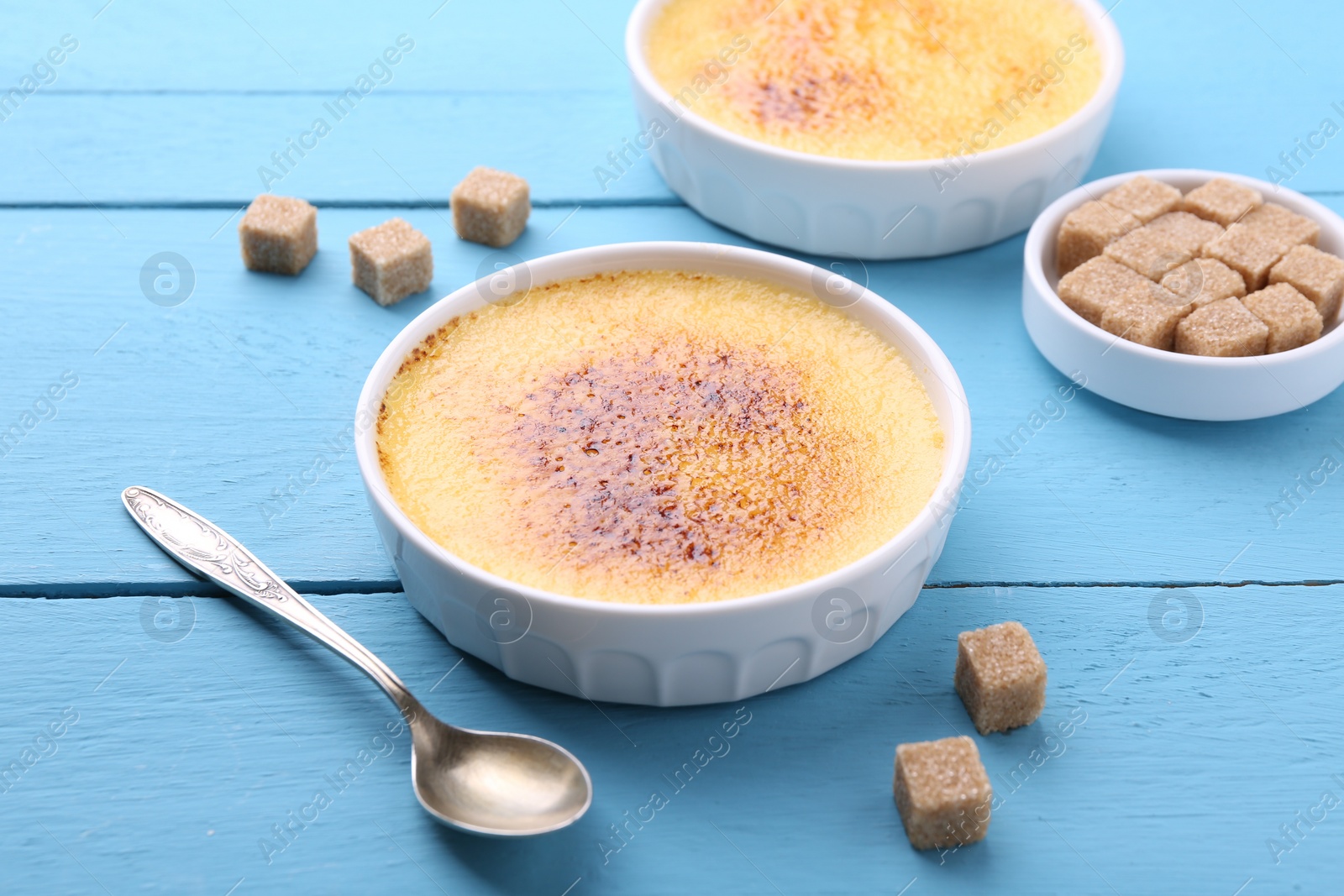 Photo of Delicious creme brulee in bowls, sugar cubes and spoon on light blue wooden table